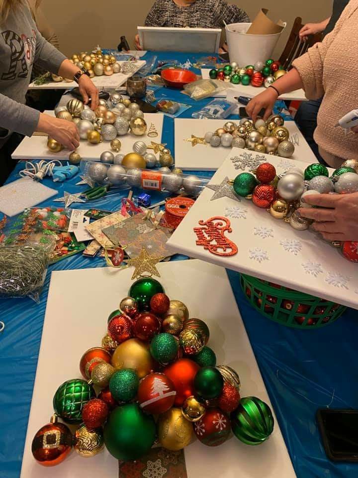 People-gathered-around-a-table-creating-Christmas-tree-wall-art-using-colorful-ornaments,-canvas-boards,-and-hot-glue.
