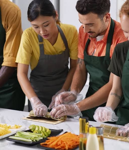 A-group-of-people-wearing-aprons-and-disposable-gloves-learning-how-to-roll-sushi-together-using-bamboo-mats.