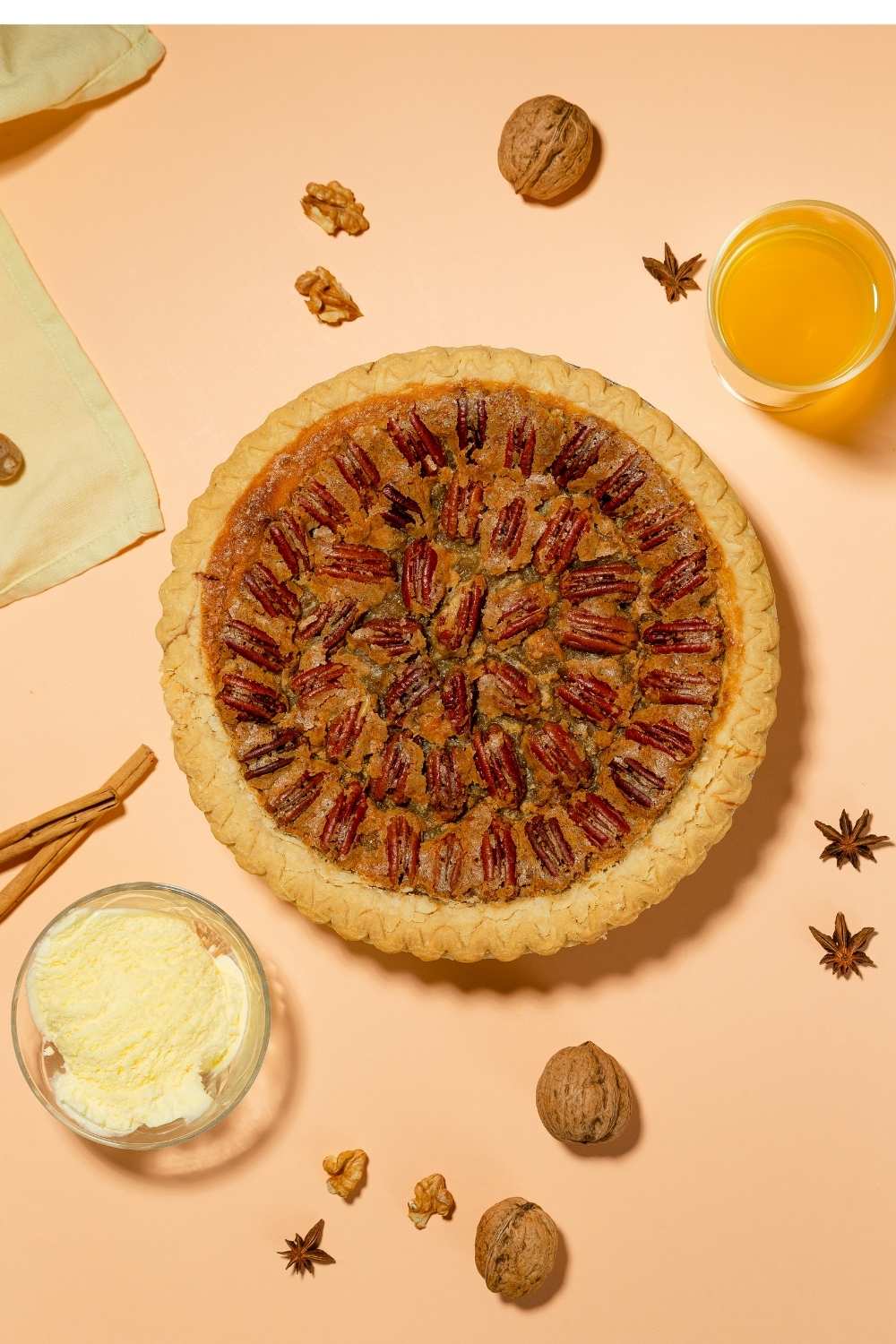 overhead-view-of-a-whole-baked-pecan-pie-surrounded-by-vanilla-ice-cream-walnuts-cinnamon-sticks-and-star-anise-on-a-peach-background