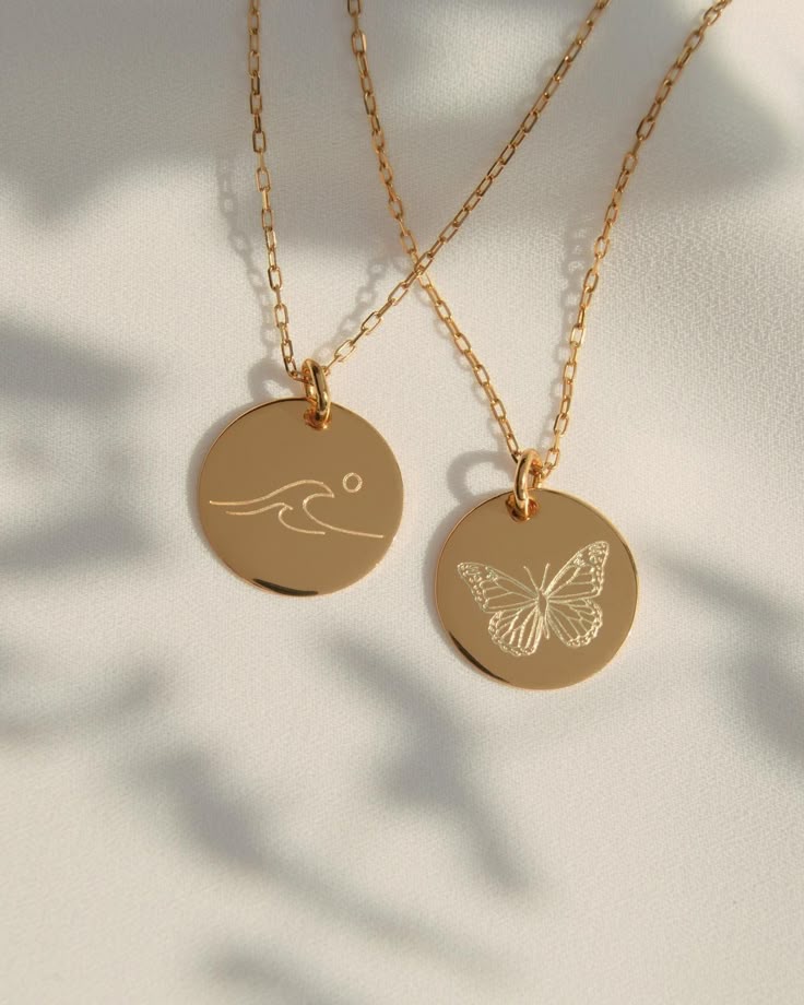 Two-gold-circular-pendant-necklaces-side-by-side;-one-engraved-with-a-wave-design,-the-other-with-a-butterfly.