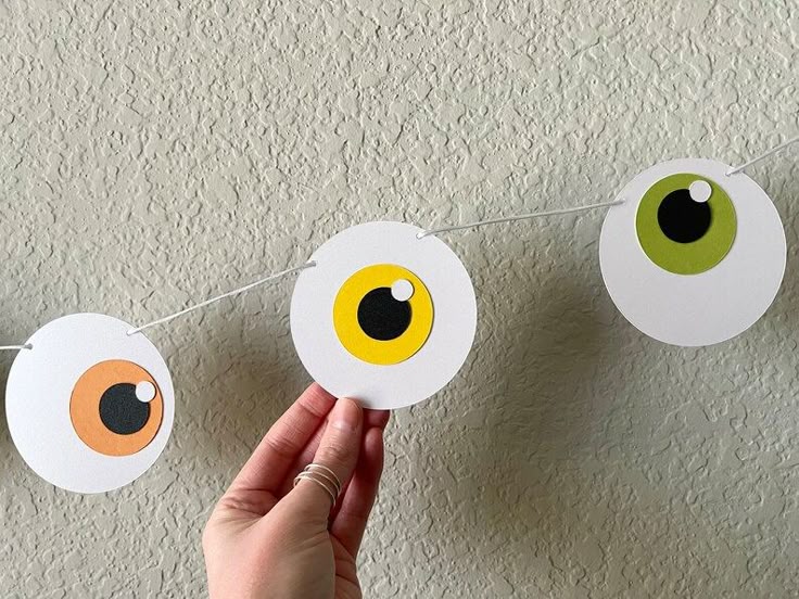 diy-halloween-paper-eyeball-garland-held-against-a-textured-wall-showing-colorful-orange-yellow-and-green-eyeballs