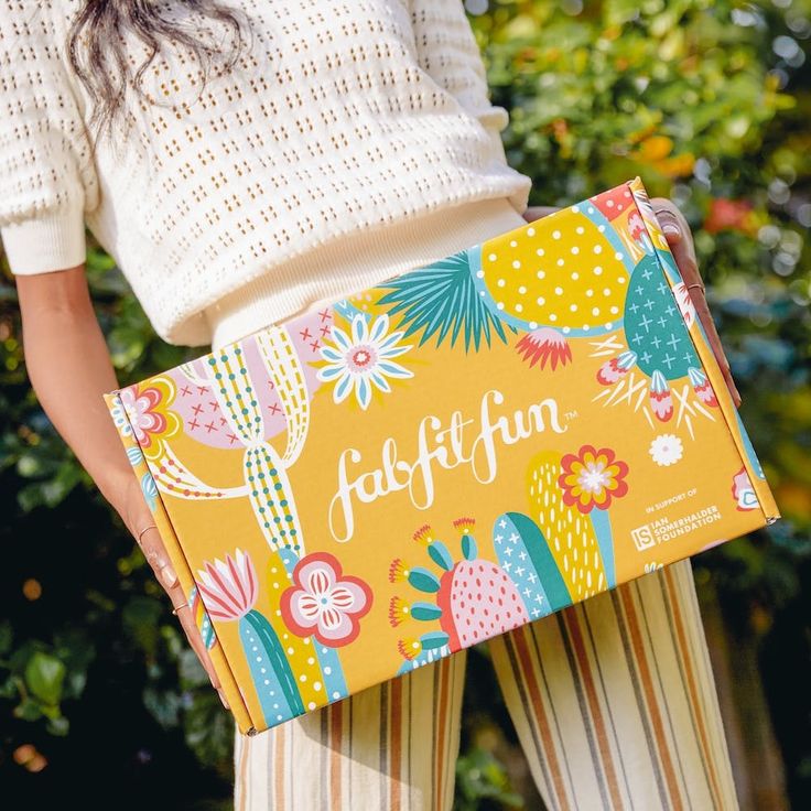 A-woman-holding-a-bright-yellow-FabFitFun-subscription-box-decorated-with-colorful-cactus-and-flower-illustrations.