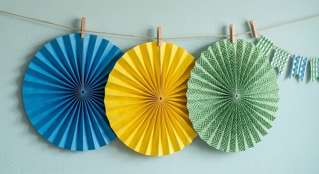 colorful-accordion-paper-fans-hanging-on-string-garland-with-wooden-clothespins-against-light-blue-wall-party-decor